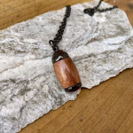 Angled top-down view of an extra small Hearthwood urn pendant made from Hawaiian Koa wood with polished black metal end caps, resting on a stone surface and attached to a black chain for holding cremation ashes.