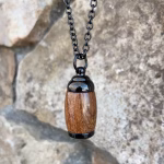 Straight-on view of an extra small hearthwood urn pendant made from Hawaiian Koa wood with polished black metal end caps, suspended from a black chain for holding cremation ashes.