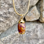 Straight-on front view of an extra small royal honey acrylic urn pendant with a gold-colored stainless steel core, suspended from a gold snake chain against a stone background for holding cremation ashes.