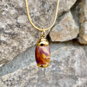 Straight-on front view of an extra small royal honey acrylic urn pendant with a gold-colored stainless steel core, suspended from a gold snake chain against a stone background for holding cremation ashes.