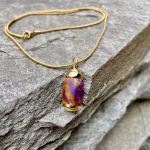 Angled top-down view of an extra small royal honey acrylic urn pendant with a gold-colored stainless steel core, resting on a stacked stone surface and attached to a gold snake chain for holding cremation ashes.