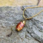 Angled side view of an extra small royal honey acrylic urn pendant with a gold-colored stainless steel core, resting on a stone surface and attached to a gold snake chain for holding cremation ashes.