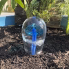 Glass jellyfish cremation paperweight displayed outdoors in soil with flowing blue glass tentacles and cremation ash suspended in clear glass