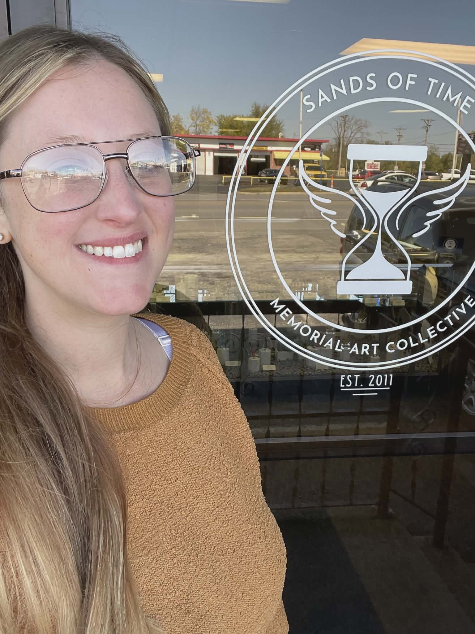Smiling Sands of Time team member outside the storefront entrance with Sands of Time Memorial Art Collective logo on the glass door