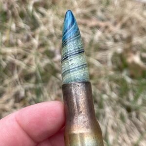 A memorial glass bullet tip in blue with white cremation ash set inside a brass casing held in hand outdoors.